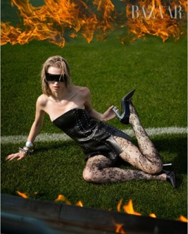 Model posing on grass in a black dress with flames in the background.
