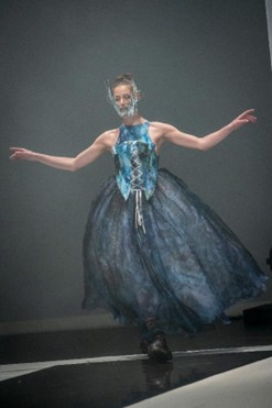 Runway model in a blue structured top and voluminous skirt with a sculptural face accessory.
