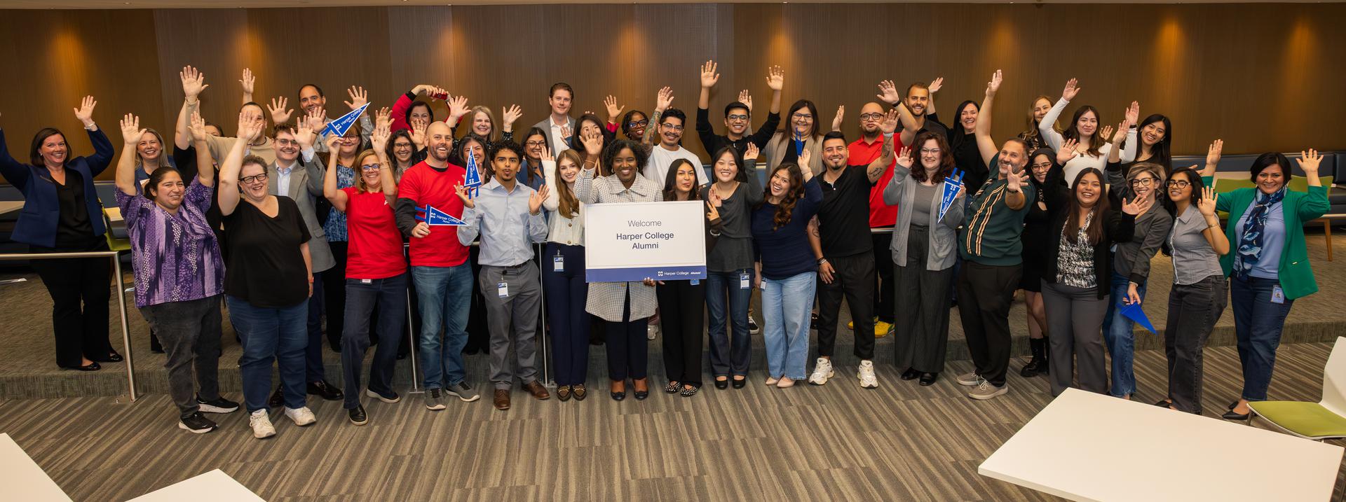 A large group of Harper College alumni and staff smiling and waving together during the Harper @ Work event at the Zurich Center.