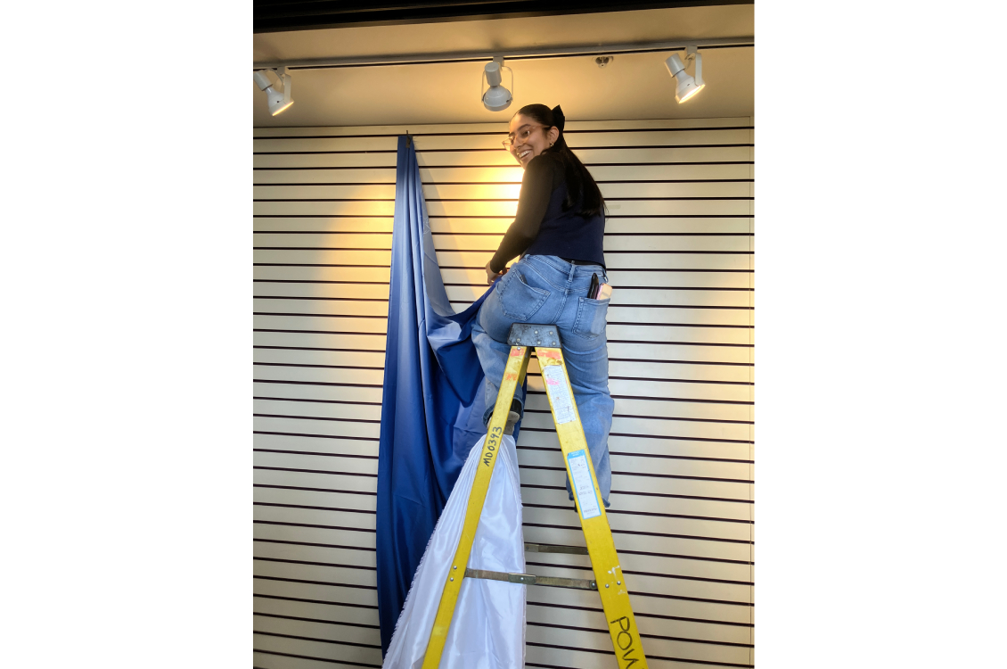 A student seated at the top of a ladder to hang up a backdrop in the display window