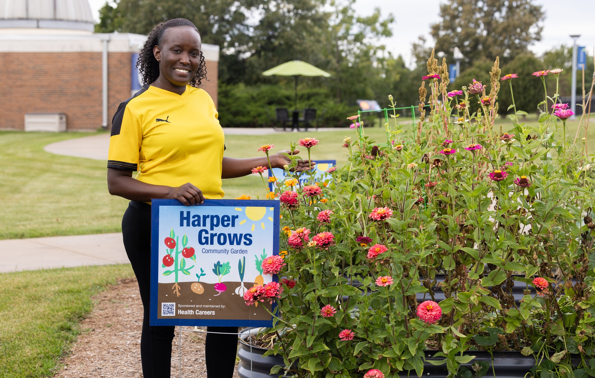 Edith Mugisha stands next to plants and a sign at the Harper Grows Community Garden on the college's campus