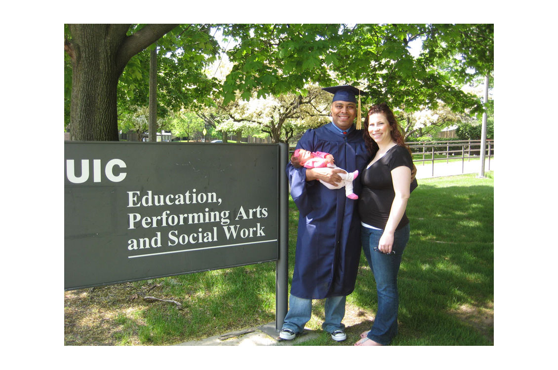 Harty in a graduation cap and gown holding a baby with his wife at graduation
