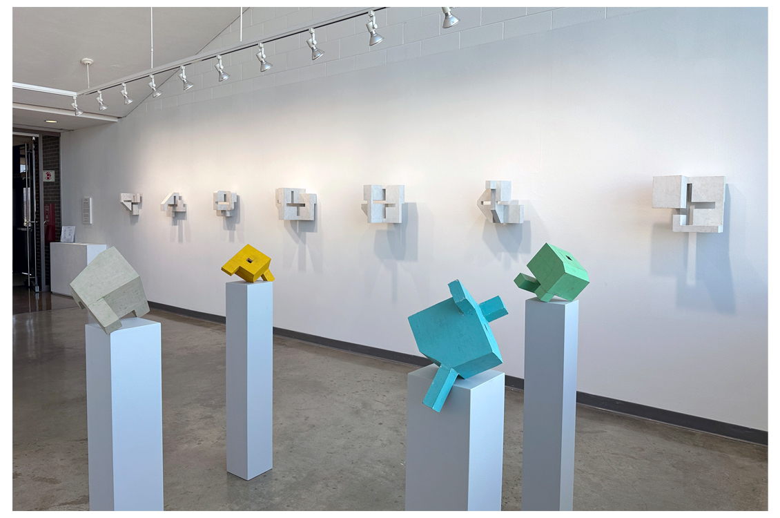 Installation view with colorful wall pieces and pale works in background.