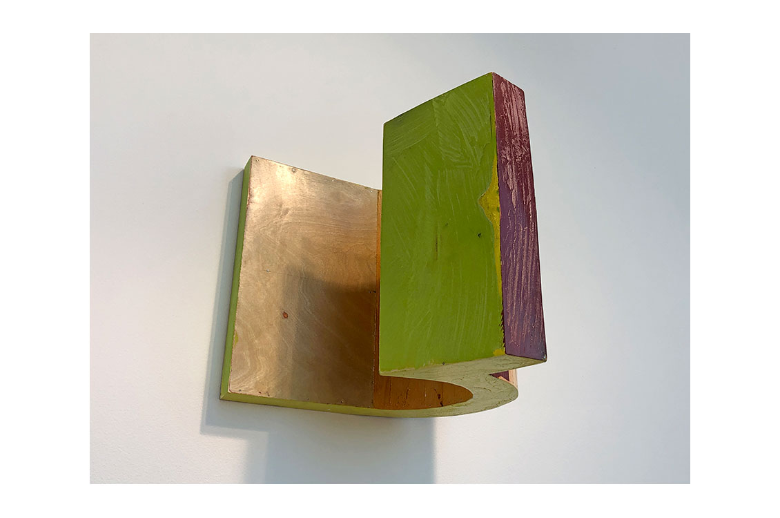 Wood sculpture hung on wall, one edge painted purple, another green.  Sculpture curves around itself on the wall.