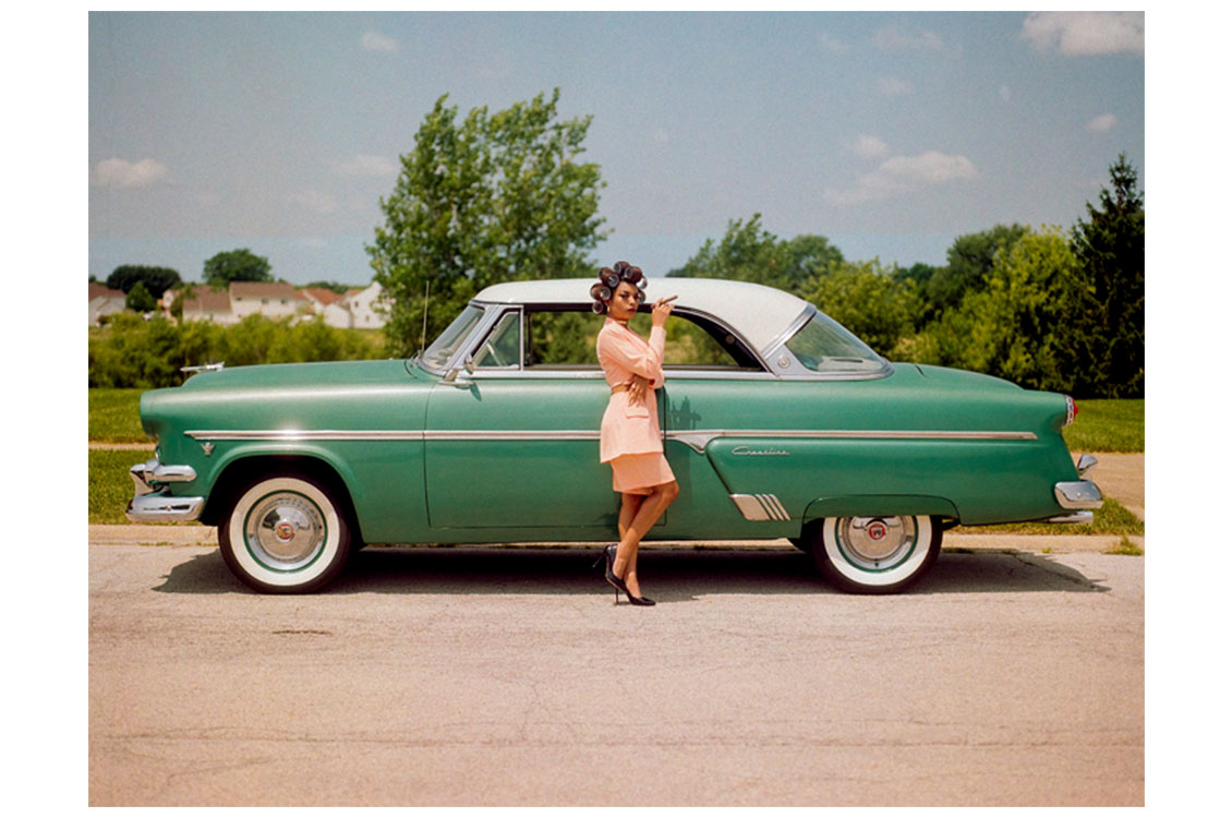 Vintage green car with white roof parked on pavement; person in peach outfit stands in front of it.