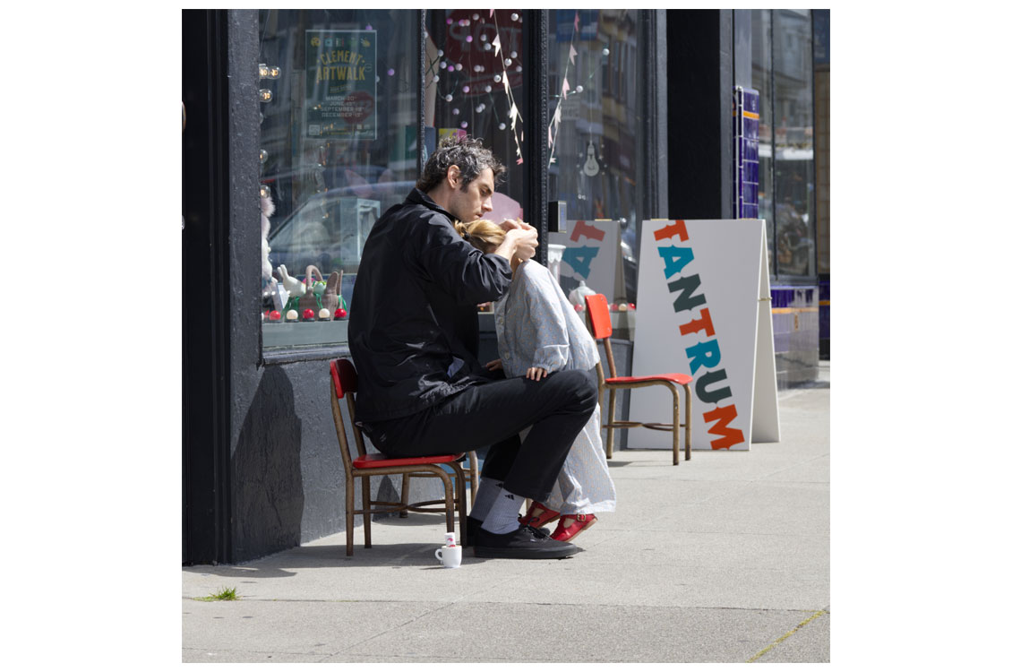 Two people sit outside a store; one adjusts the other's hair near a colorful 'TANTRUM' sign and holiday decor.