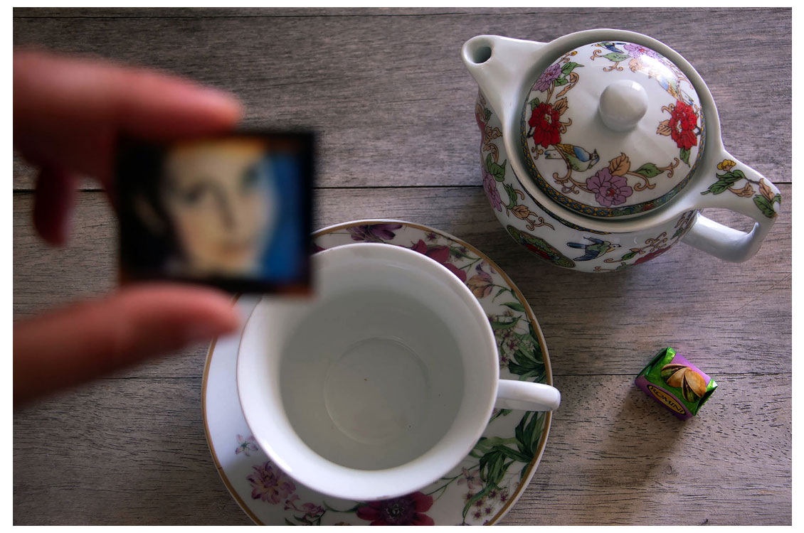 Floral teapot, matching teacup, and wrapped candy on wood surface; blurred hand holds a small photograph in foreground.