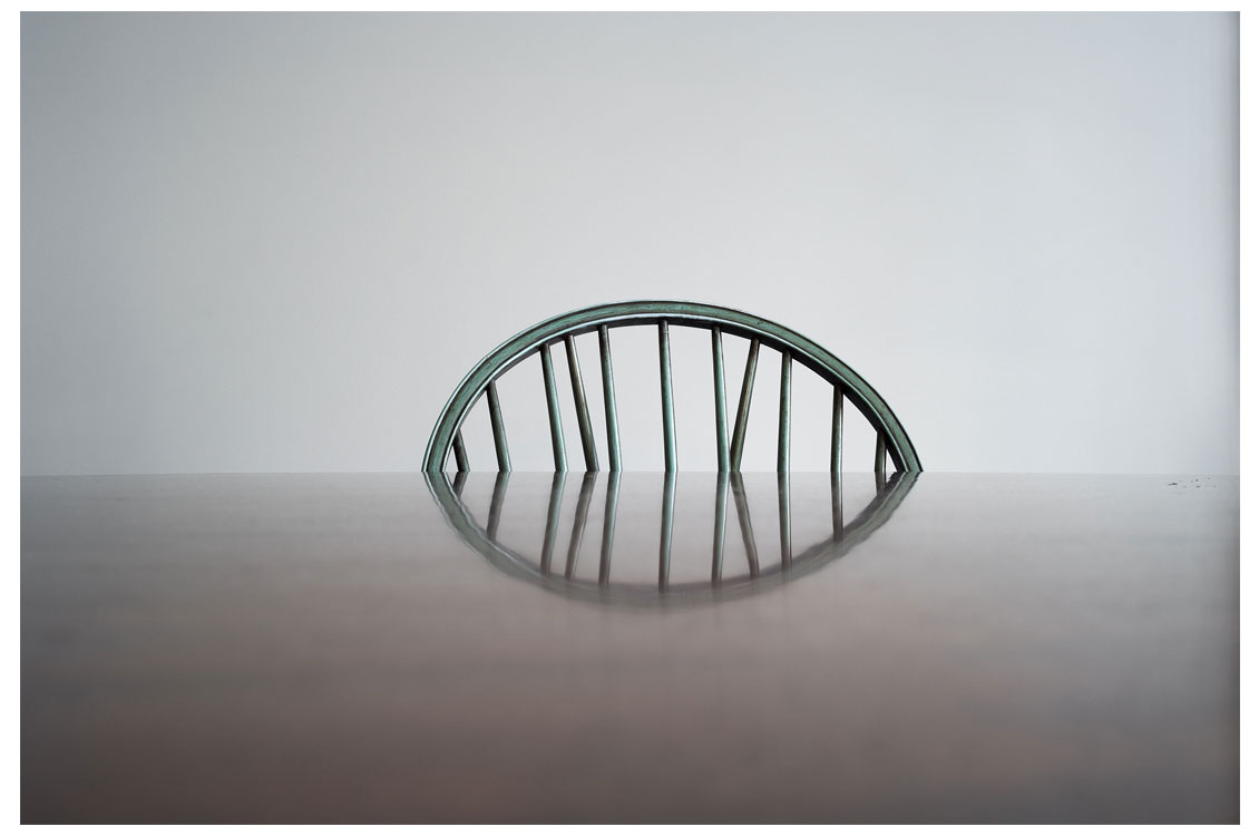 Wooden chair backrest with vertical slats reflected on a glossy table surface, set against a plain background.