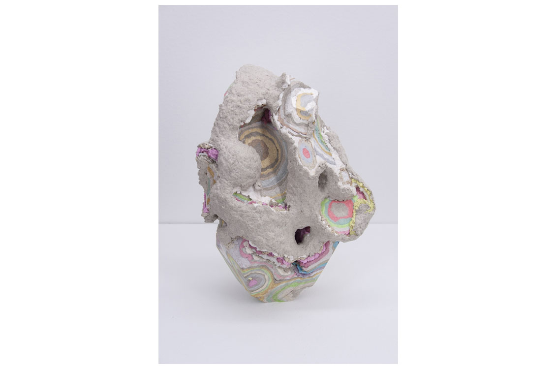 Gray sculptural object with rough surface and exposed colorful concentric layers in green, pink, yellow, and purple.