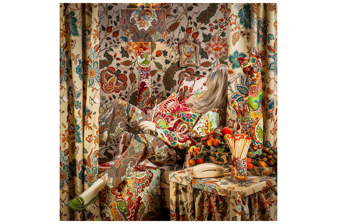 Staged photo of a person in patterned clothing reclining amid matching floral upholstery and curtains.