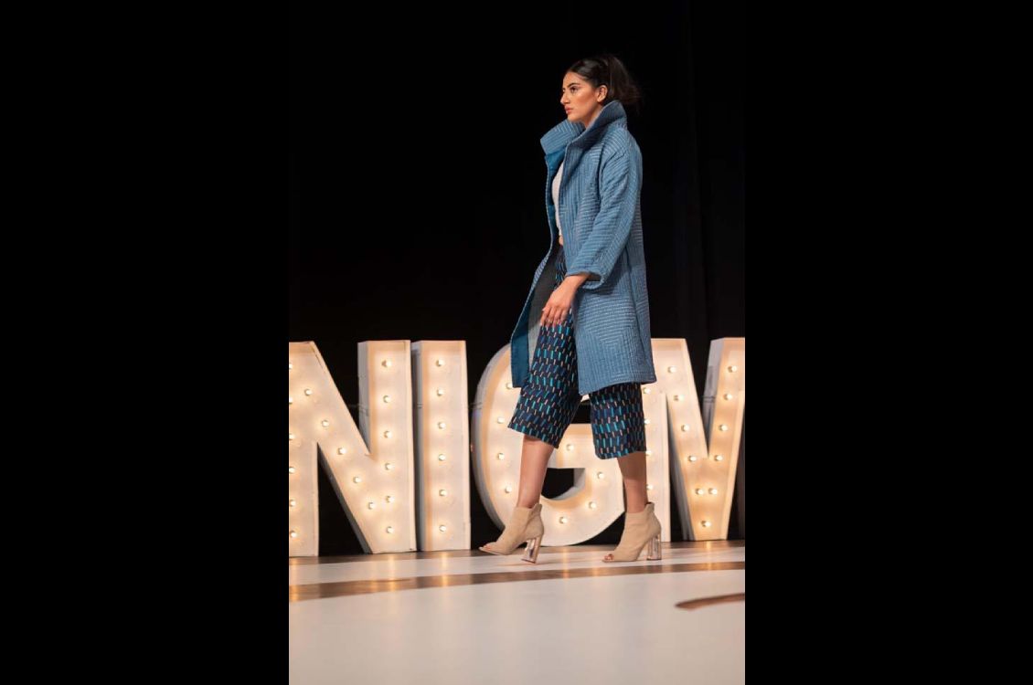 Woman walking on stage with a blue and beige ensemble under a long teal overcoat