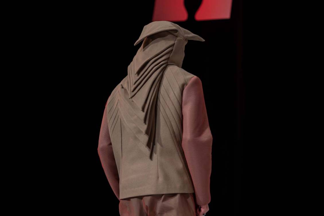The back of a male model wearing an angular brown and red hooded outfit