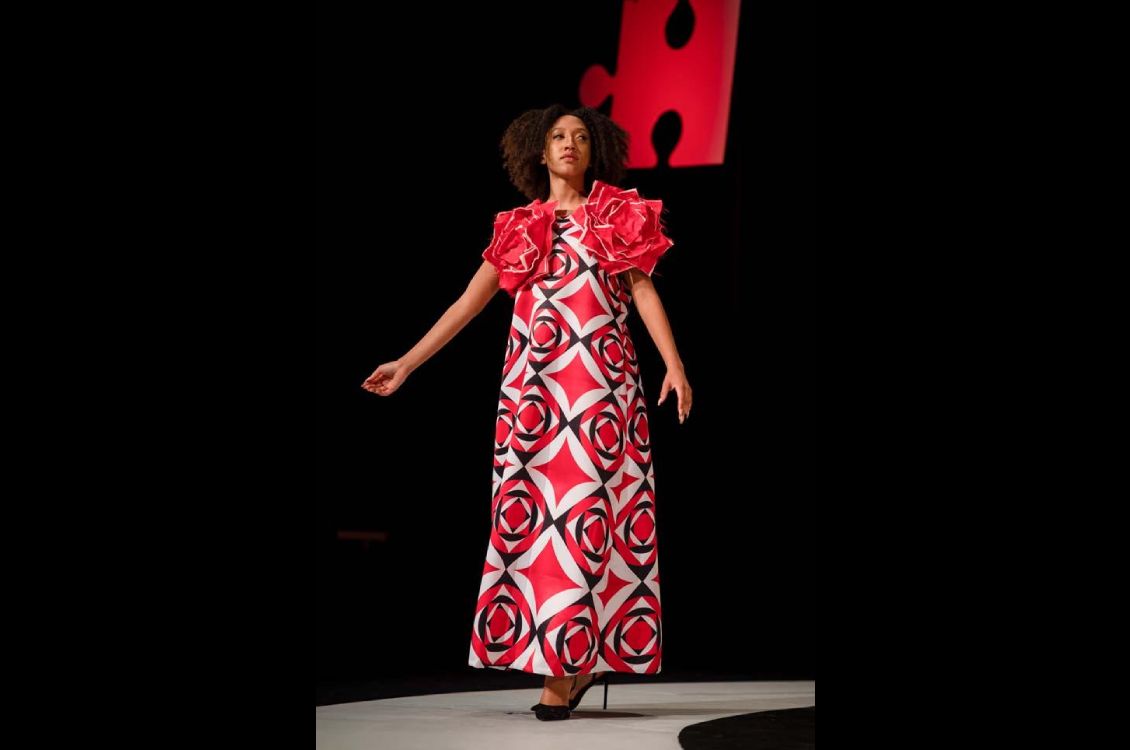 Model wearing a rong red, white, and black patterned dress with large fabric rosettes on the shoulders