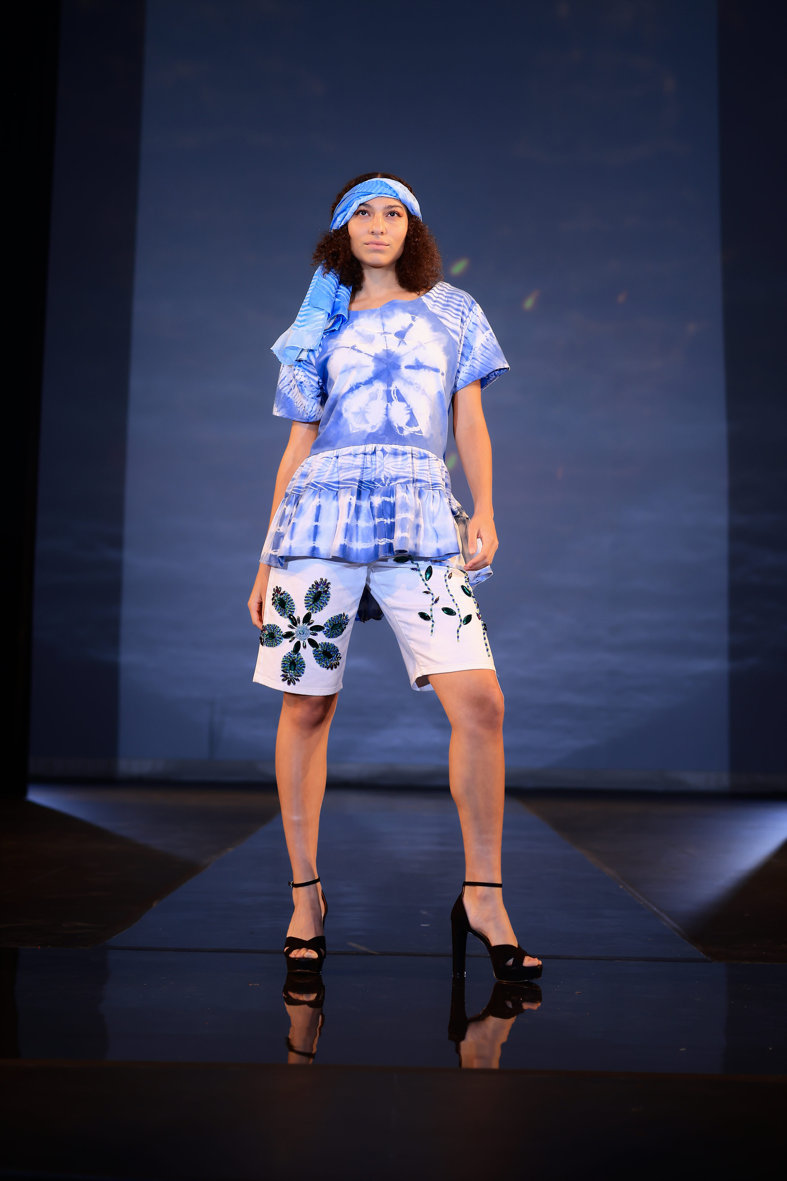 Model is wearing a blue tie-dyed top, white Bermuda shorts with a black floral pattern, black heels, and a blue tie-dyed scarf on her head.