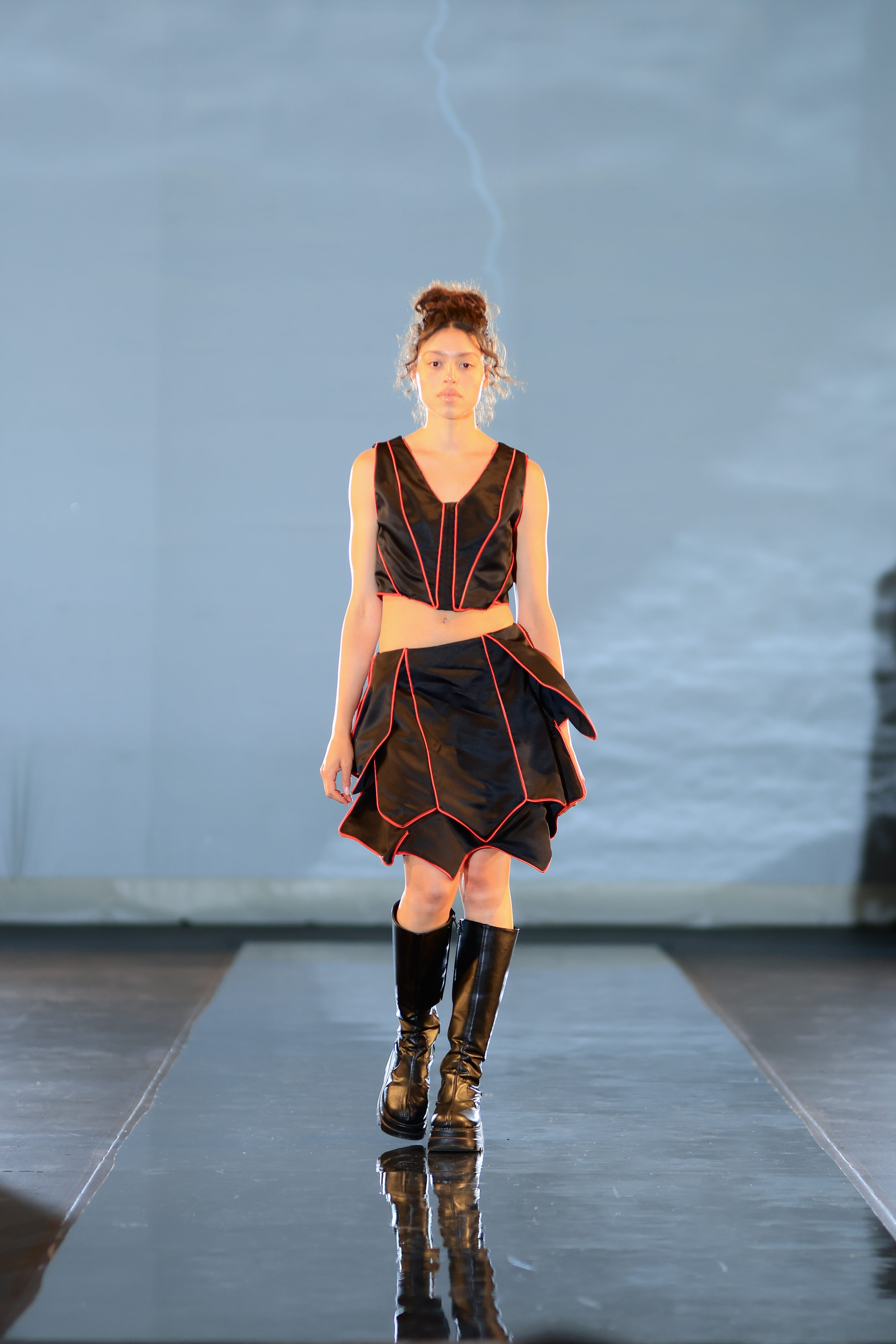 A model is strutting down the runway in a black and red two-piece dress paired with black boots.