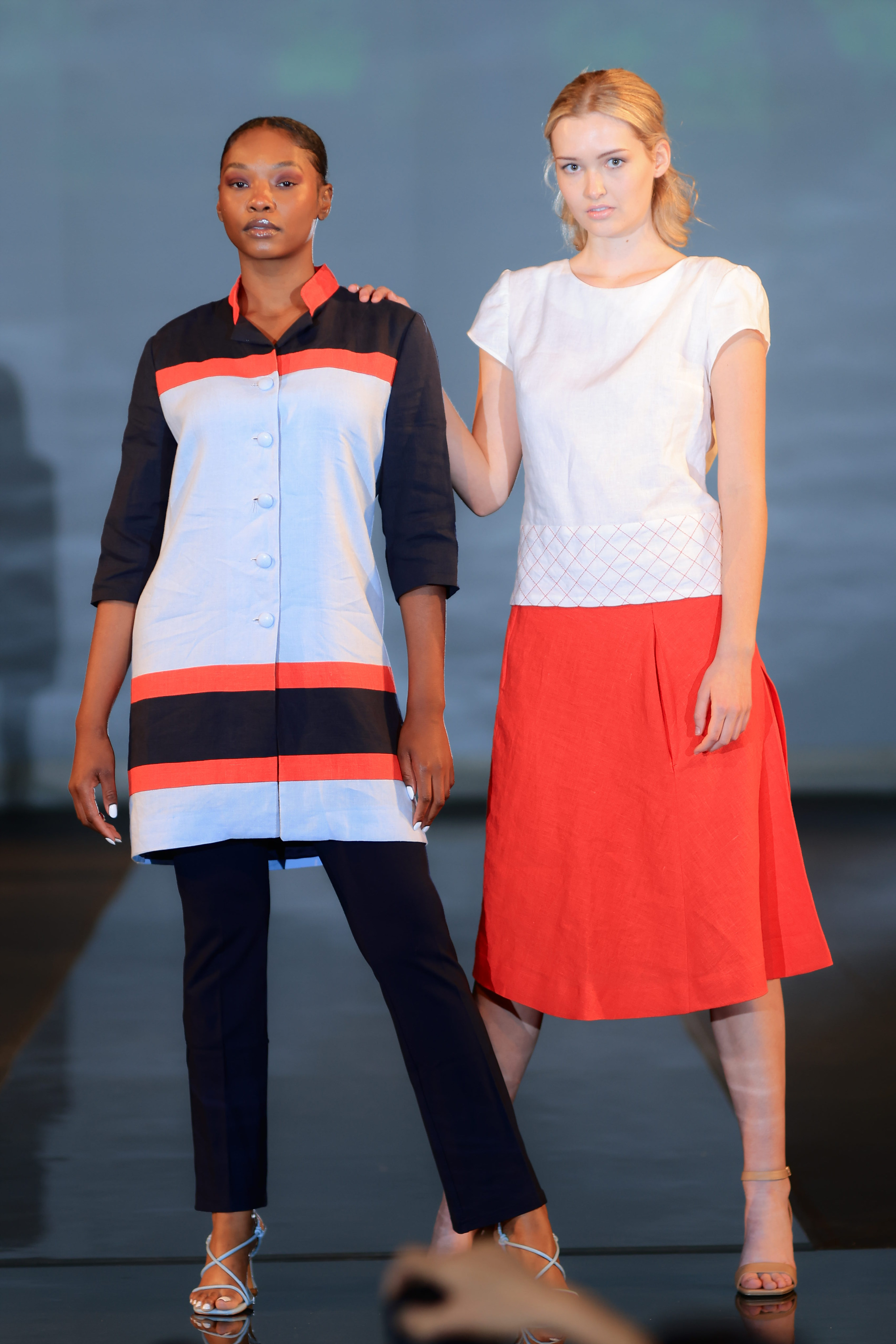 At the runway's end, two models strike a pose. The left model dons a long-sleeved black and red striped top, paired with black slacks and blue heels. Meanwhile, the right model showcases a white patterned top elegantly matched with a vibrant red knee-length skirt.