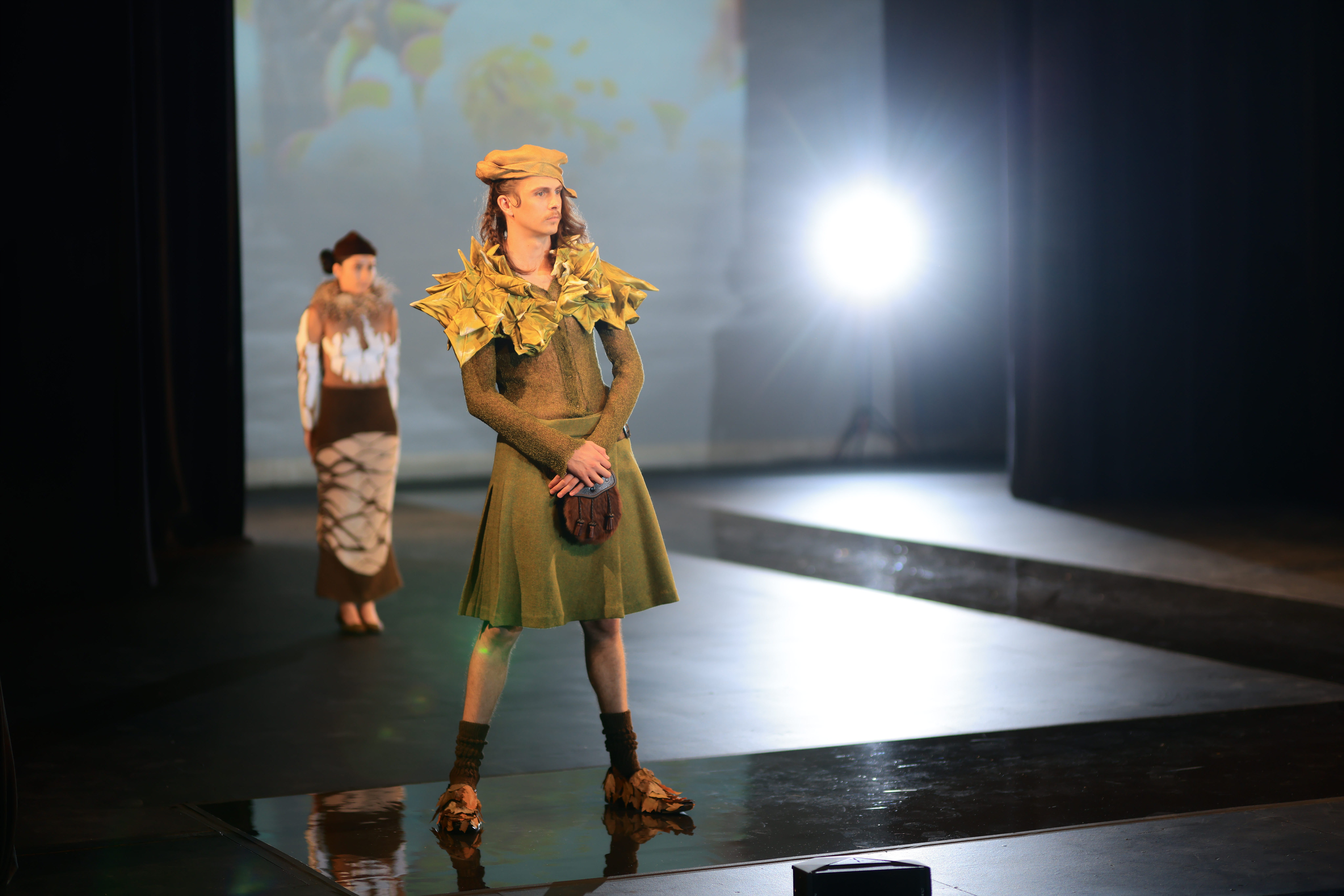 On the runway, a model strikes a pose in a vibrant ensemble, donning a green hat, a long-sleeved green top with a leafy shoulder cover, a coordinating green skirt, dark brown tube socks, and stylish loafers adorned with brown leaves. In the background, another model awaits their turn to walk.