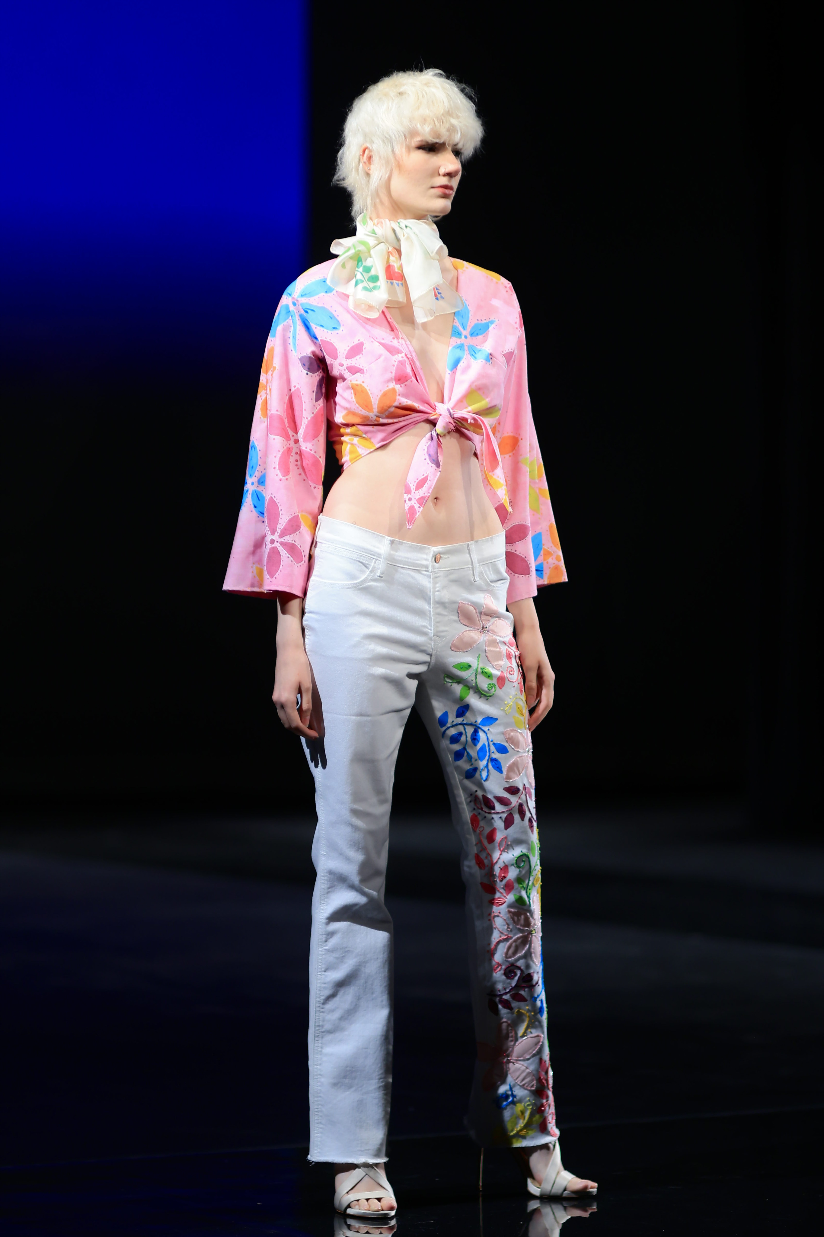A model wears a chic ensemble: a white scarf with a colorful floral pattern, a pink tied cropped top with matching floral design, light-washed denim jeans with a floral-covered left leg, and elegant silver-strapped heels.
