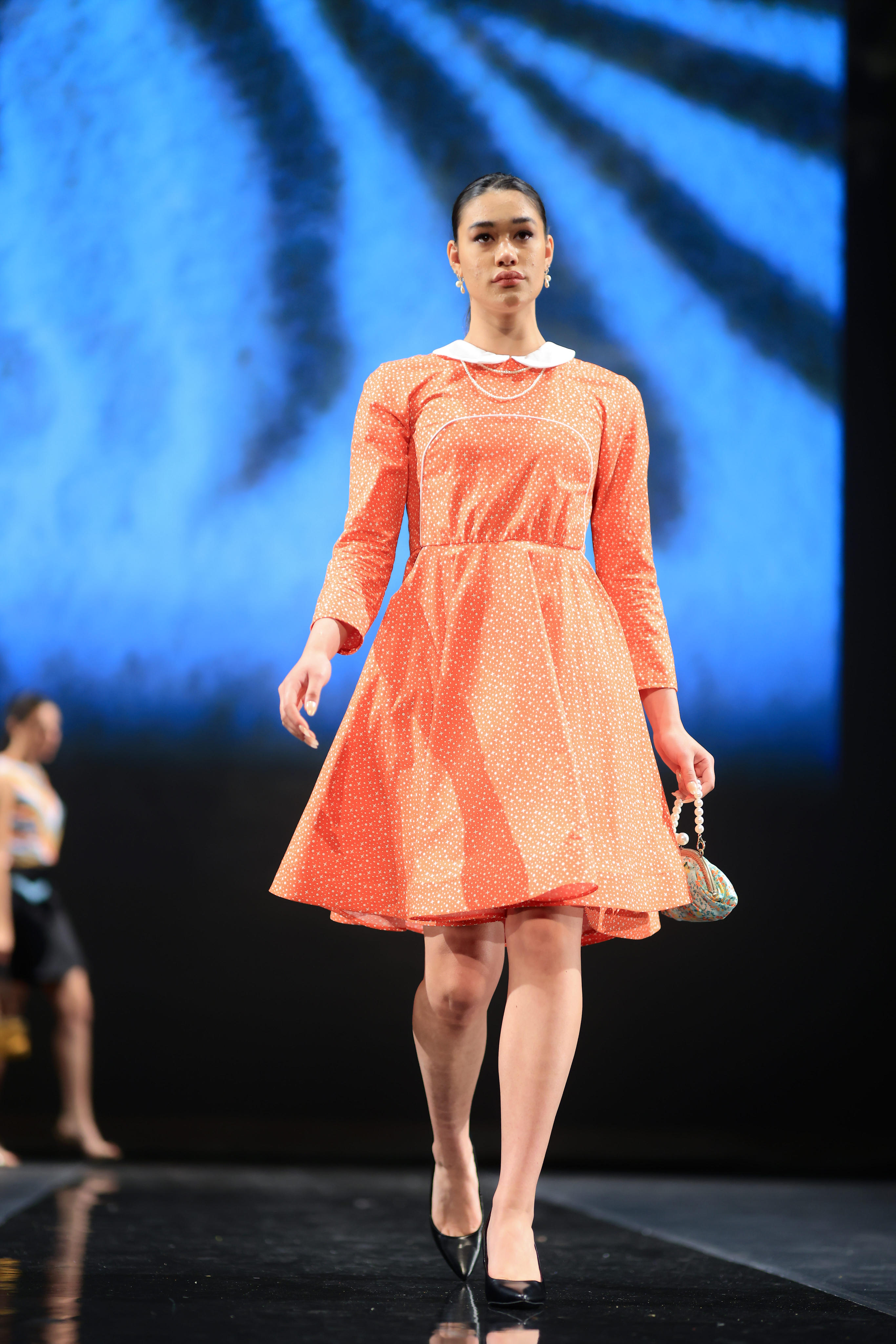 A model struts down the runway in a vibrant orange and white polka-dotted above-the-knee length dress, complemented by black heels. The ensemble is adorned with pearl earrings, and she carries a chic green and orange mini purse with pearl handles.
