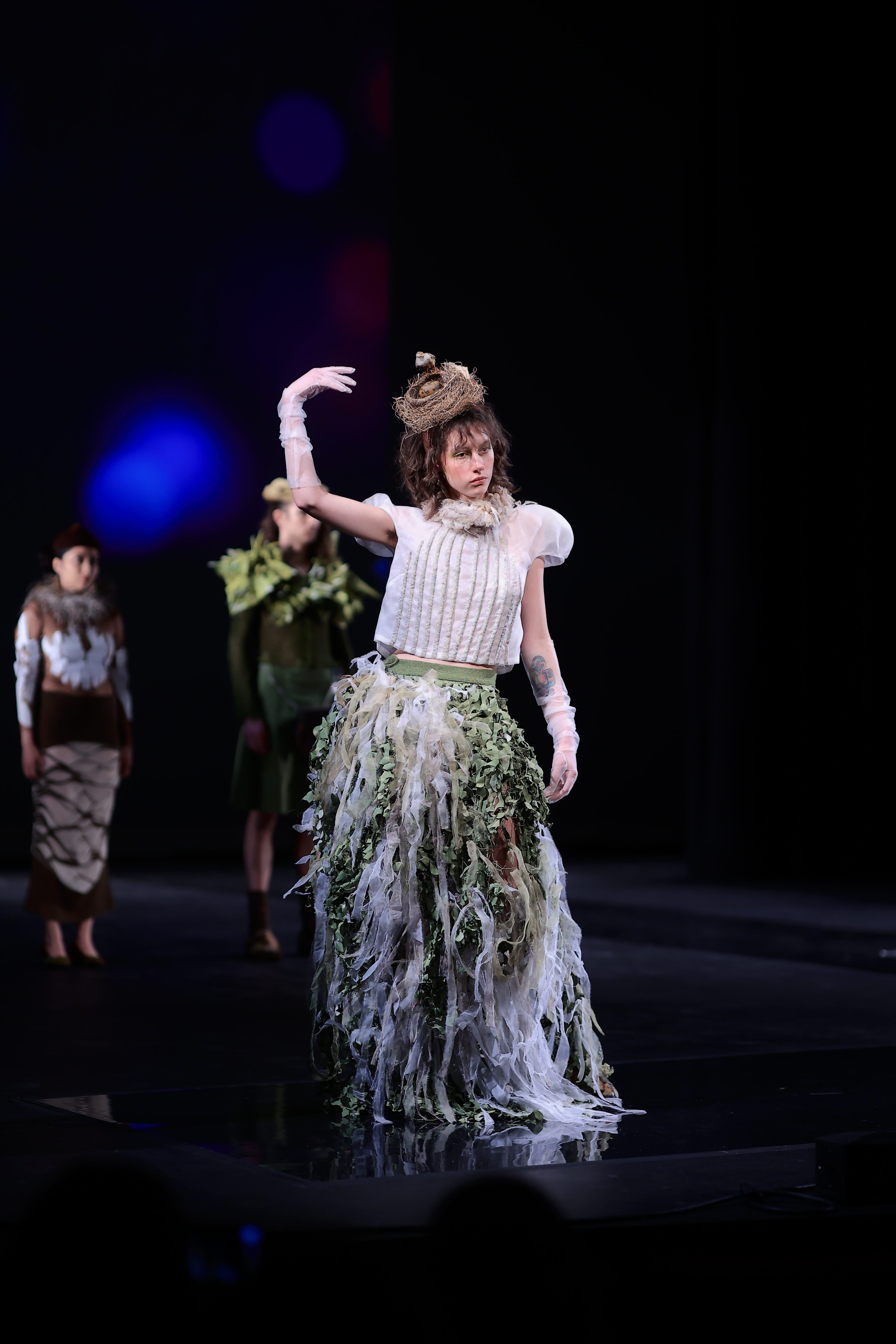 A model strikes a pose with one arm raised, showcasing a stunning ensemble: a white and green textured floor-length skirt, translucent gloves, a white top with puffy shoulders, and a unique bird's nest hat.