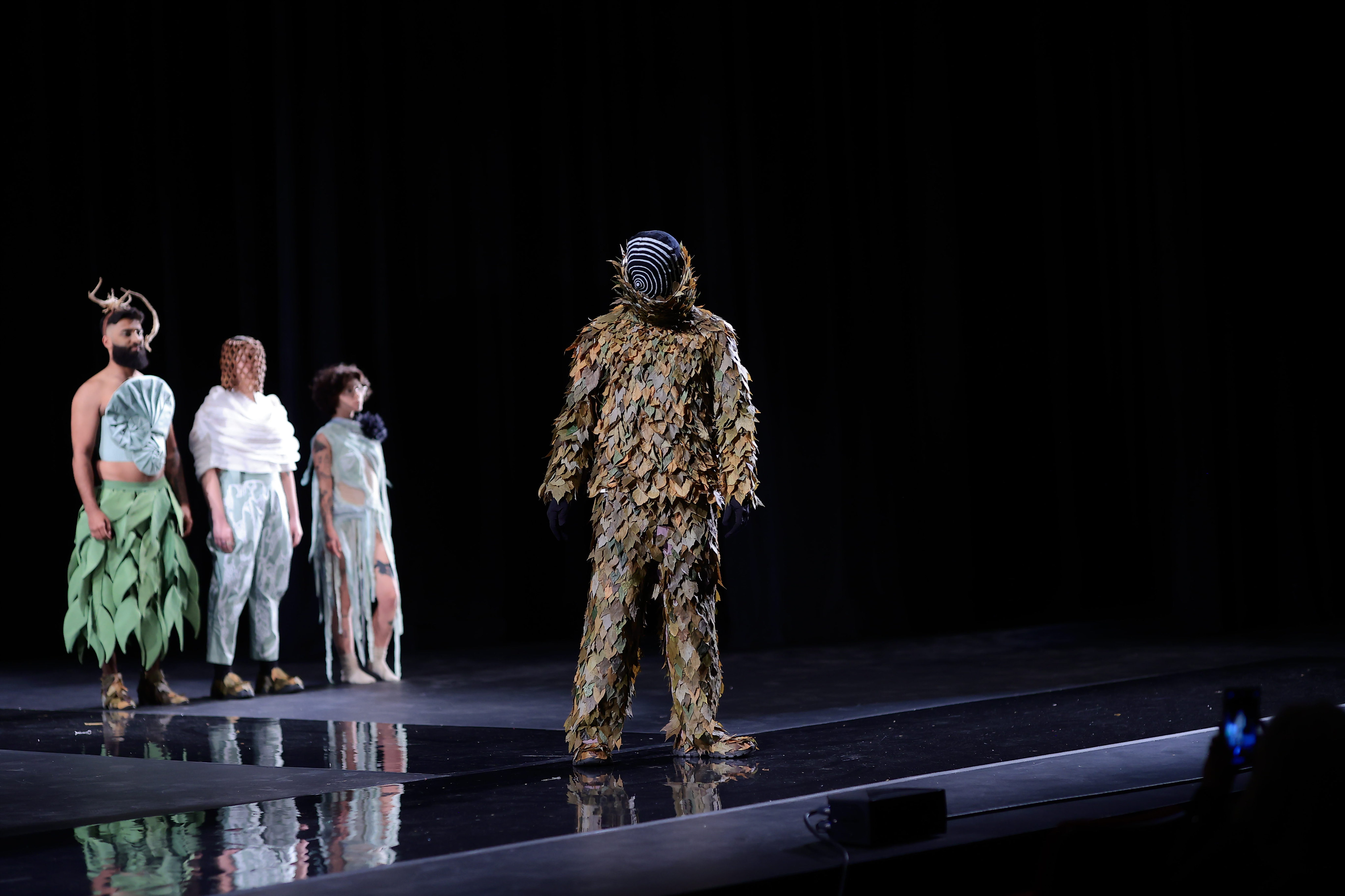 Model is wearing a leafy suit covering nearly head to toe. Models head is covered in a black mask with a white spiral on the front. Group of models pose in the background 