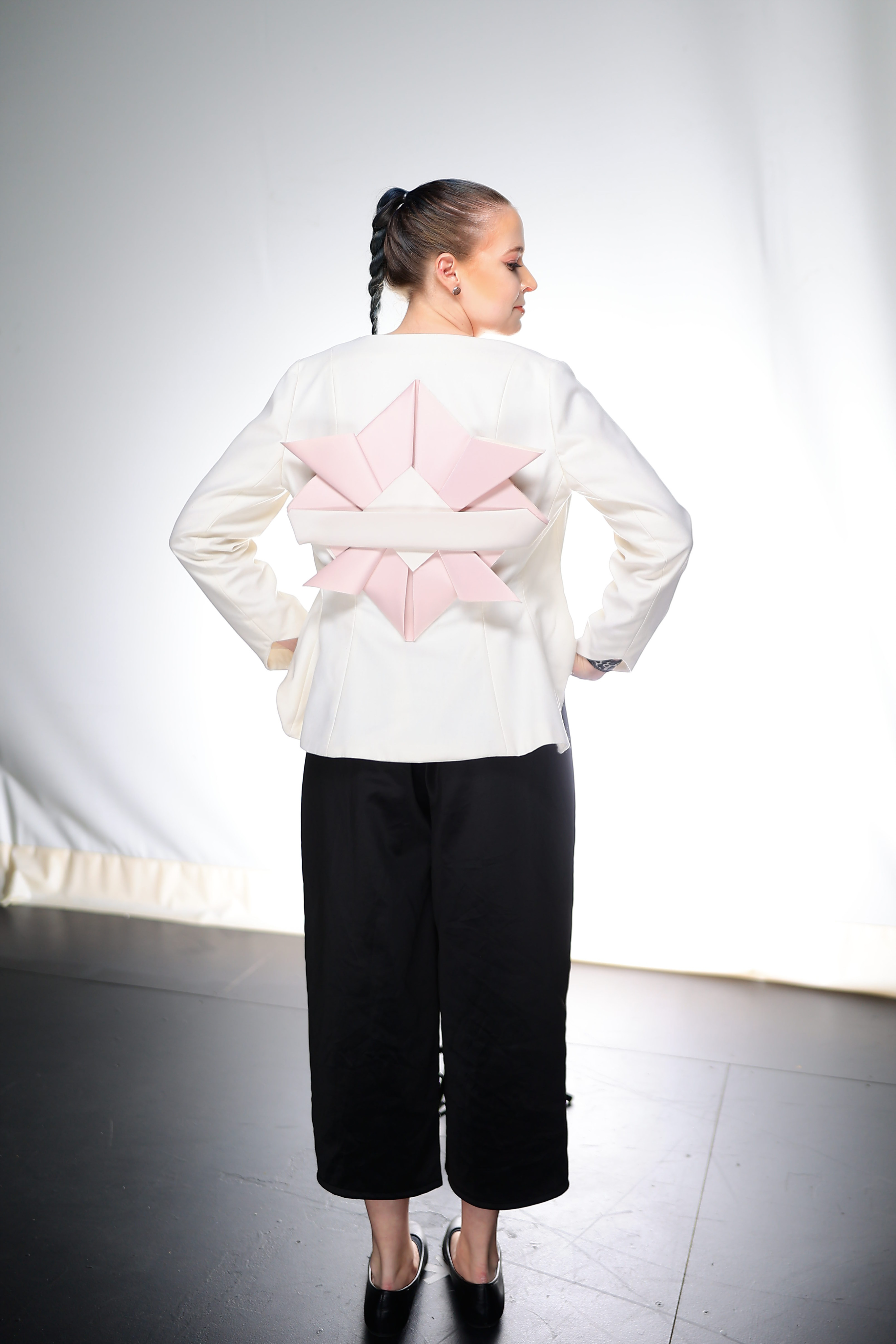 A model posing with their back to us. The model is wearing a white long-sleeved blazer with a pink and white origami design oversized black capris with black flats.