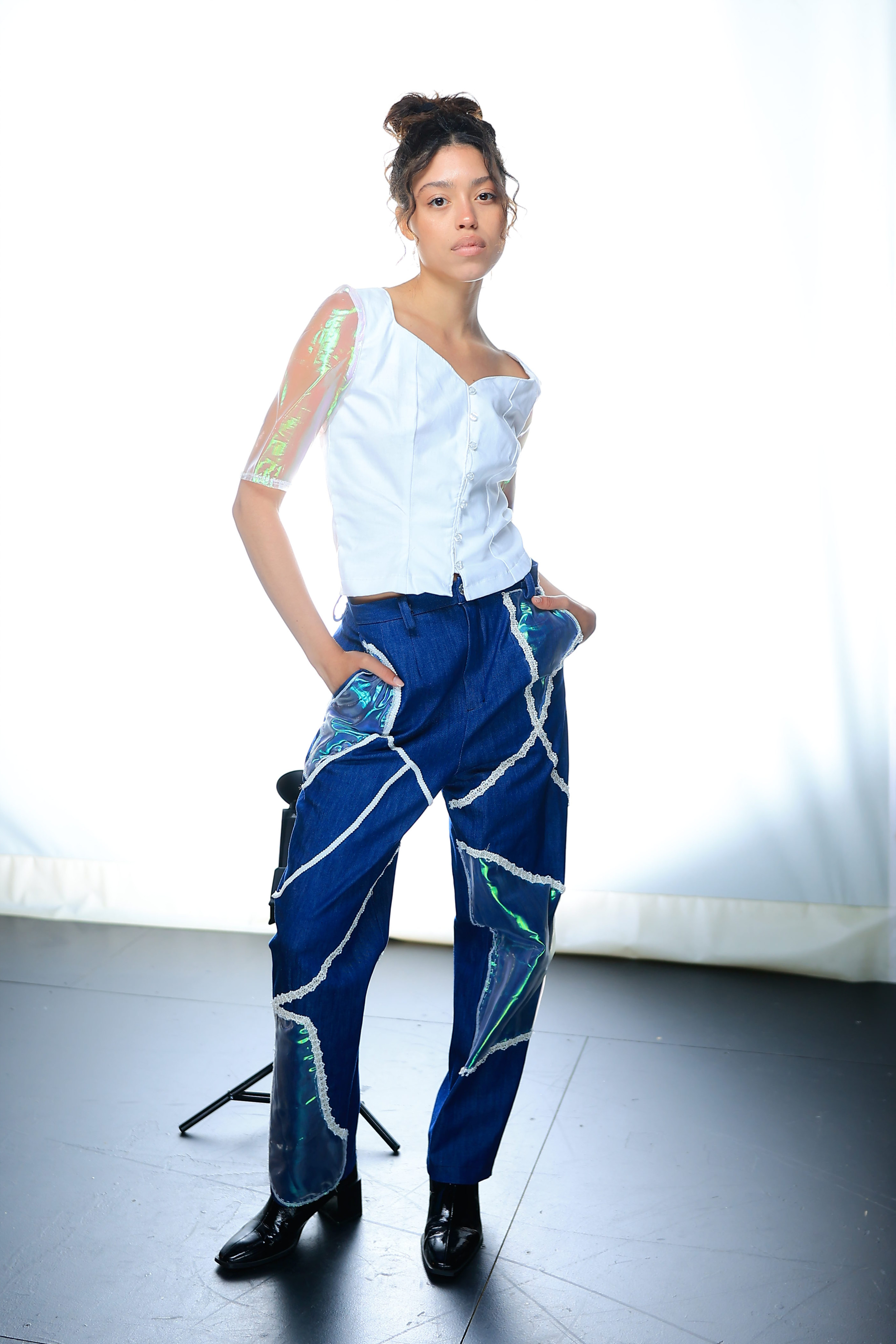 Model posing, dressed in a white top with holographic sleeves, and navy blue slacks with a holographic pattern and black heeled boots