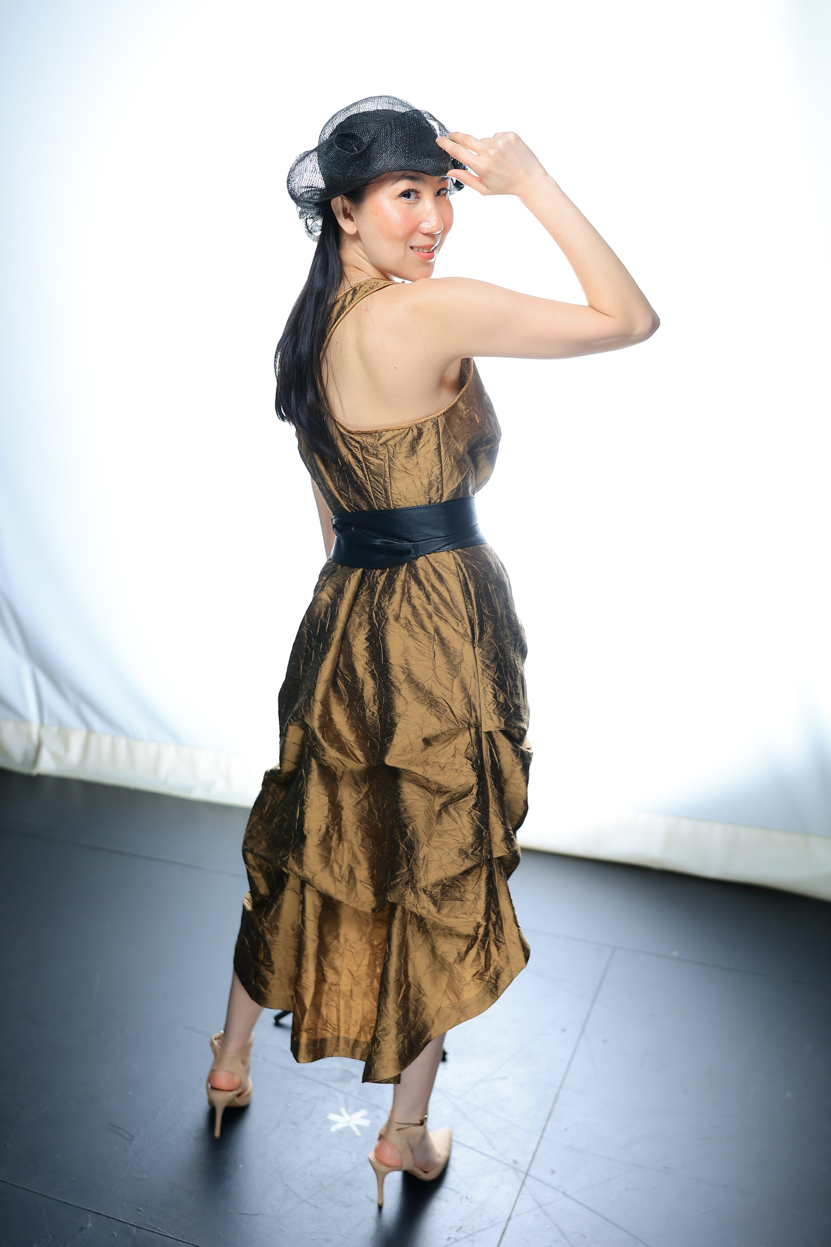 Model posed with right hand touching their black laced hat. Model is wearing beige heels and a gold and black sleeveless dress.