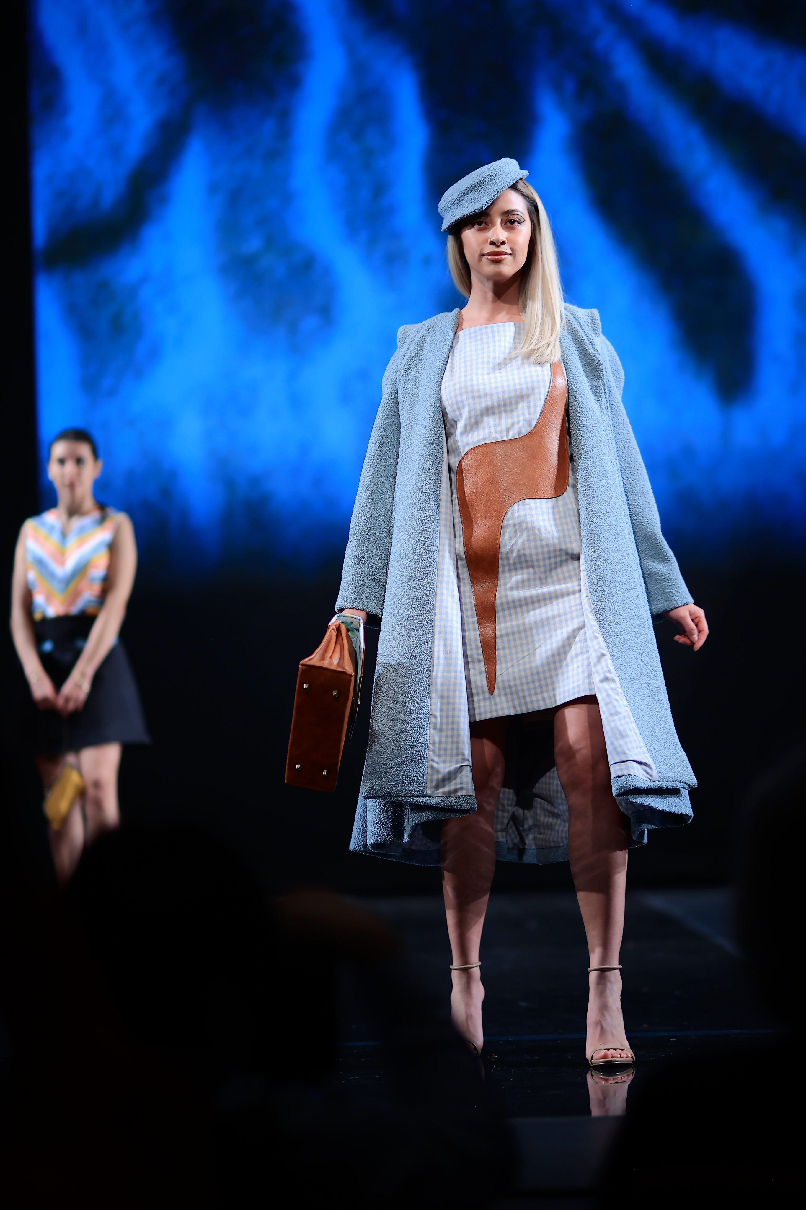 Model carrying a brown and blue purse, wearing a brown and blue patterned dress, a light blue peacoat, and a matching light blue hat.