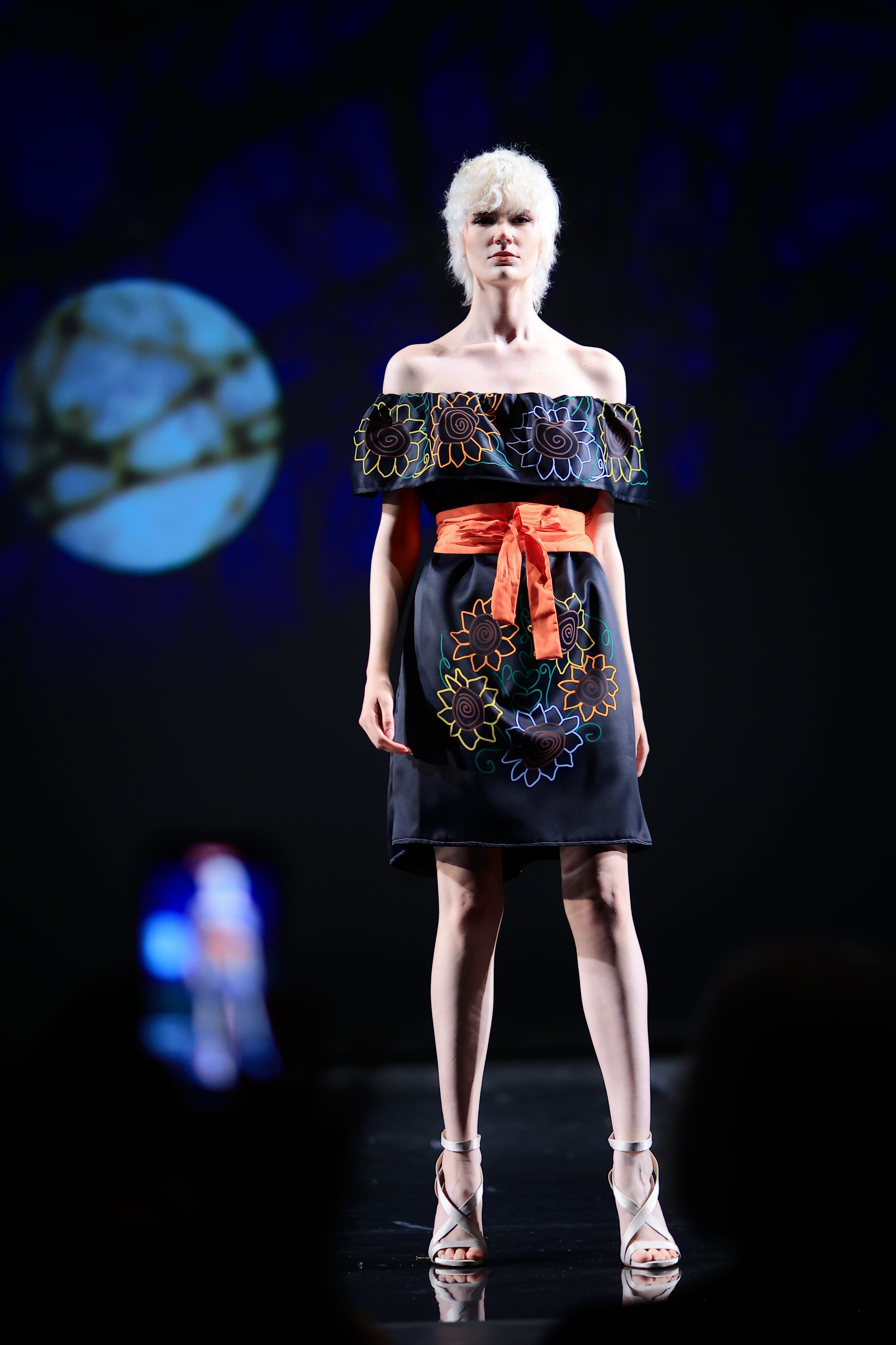 Model wearing silver strapped heels and a black dress with colorful floral pattern and an orange belt