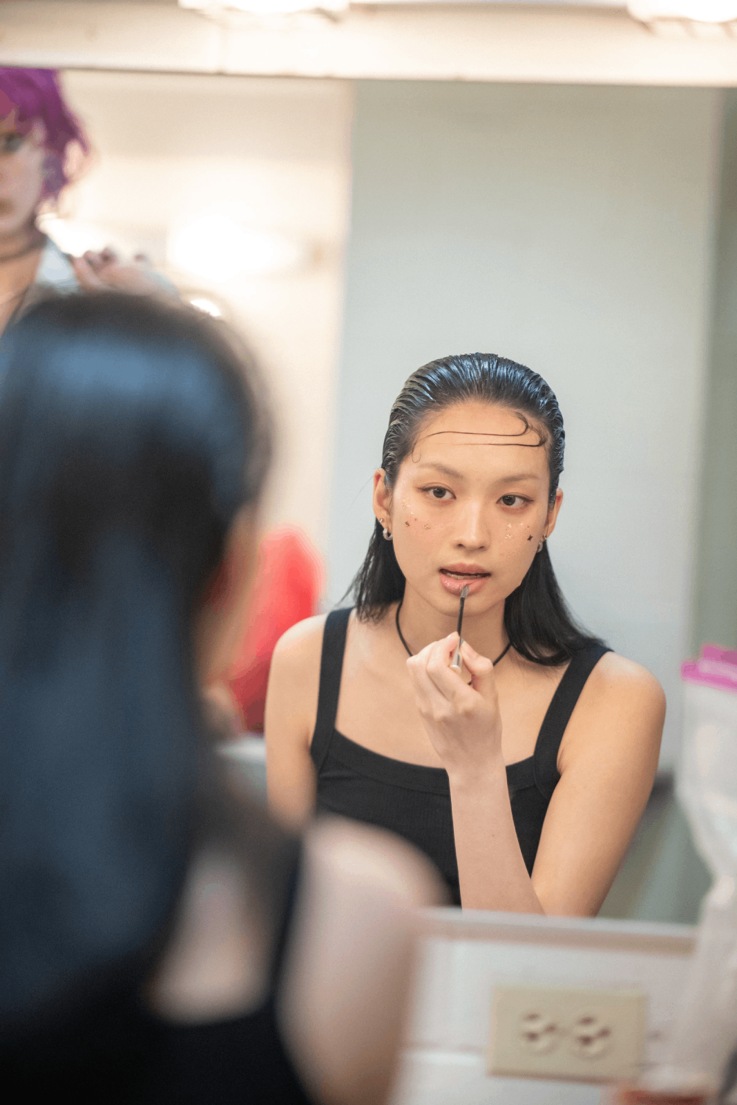 A model with long, slicked back hair applying a nude-pink lip product in the mirror backstage