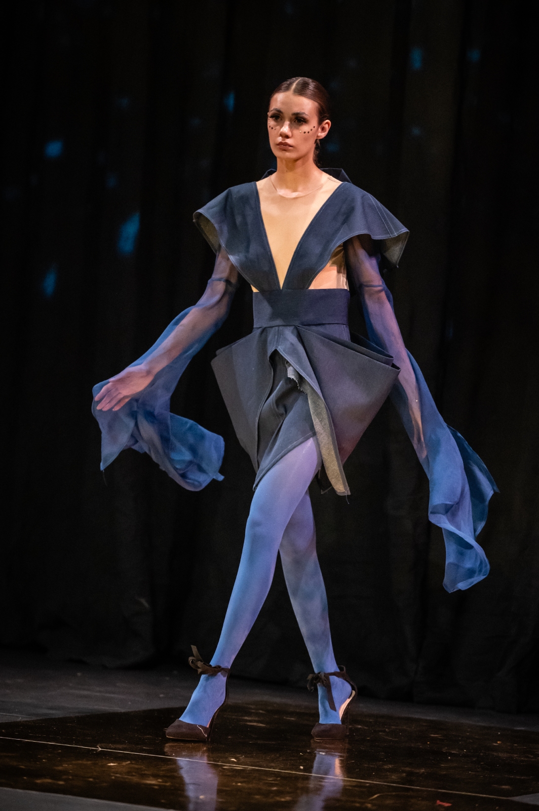 A model struts down the runway wearing a navy blue dress with cutouts at the ribs and long flowing sleeves
