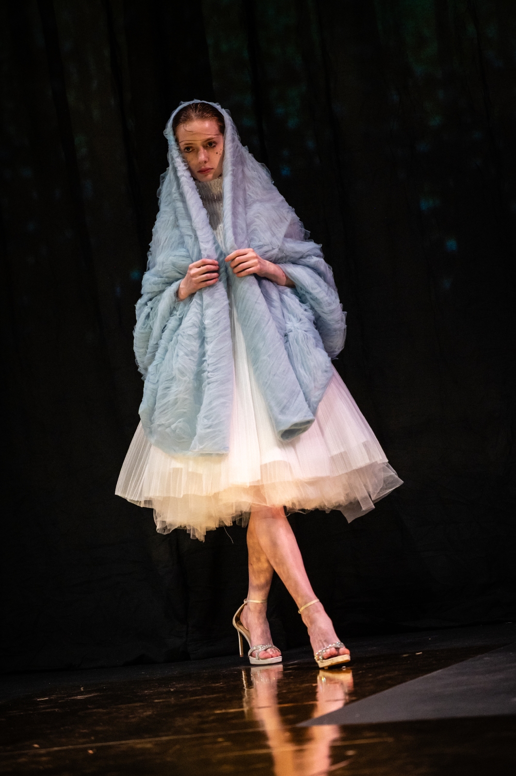 A model walks down the runway in a white, tulle ruffled skirt and a pastel blue shawl draped over her head and shoulders
