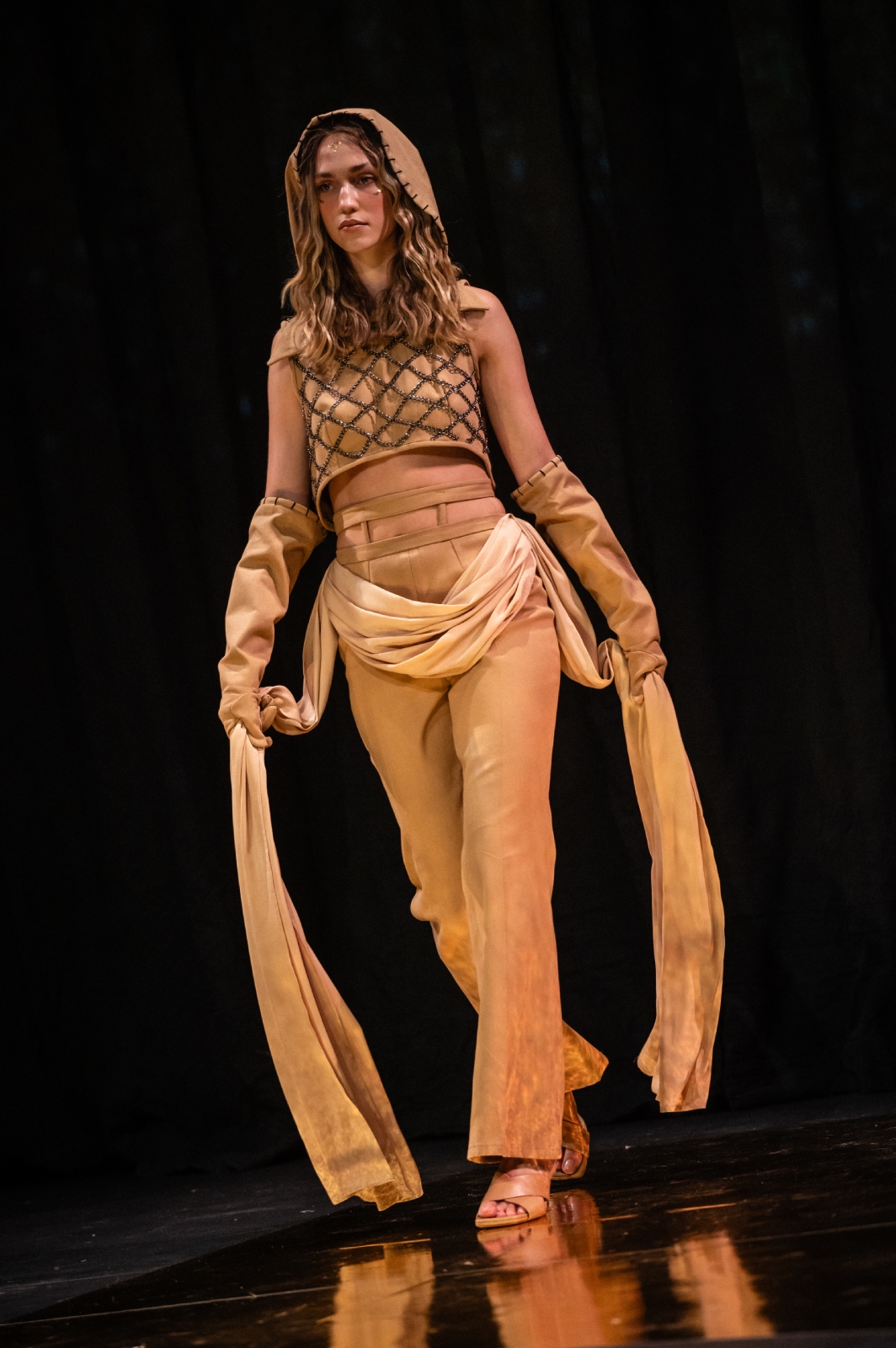 A model struts down the runway in a tan and gold outfit with a hood and long draping belt that she holds out in her hands