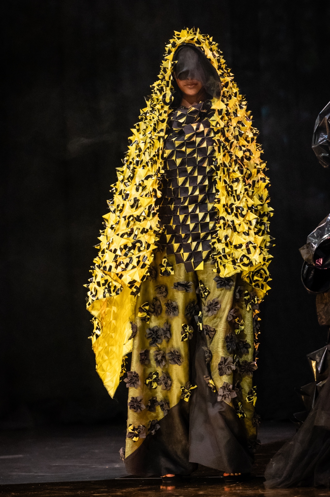A model wearing a black and chartreuse garment with a large piece of textured fabric draped over the head, and a long skirt and pants