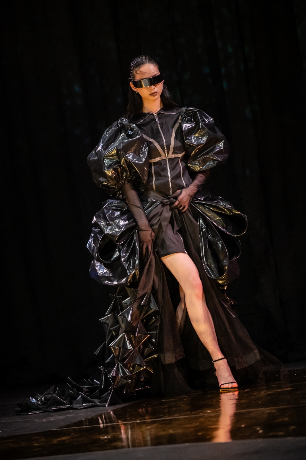 A model steps forward in a shiny black dress with a long, ruffled train and puffed sleeves