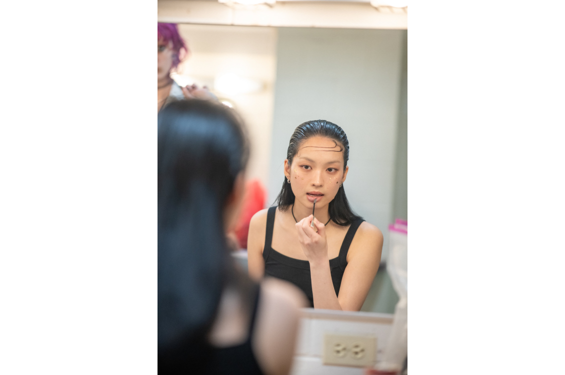 A model with long, slicked back hair applying a nude-pink lip product in the mirror backstage