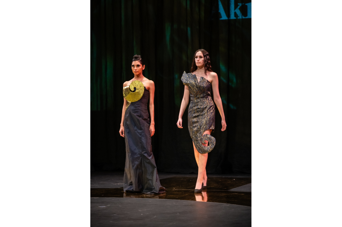 Two models join each other on the runway, wearing dark gray dresses with bright details