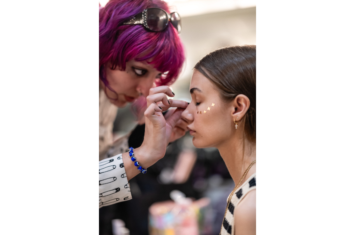A model getting gold decal sticker put on her cheekbones by a makeup artist backstage