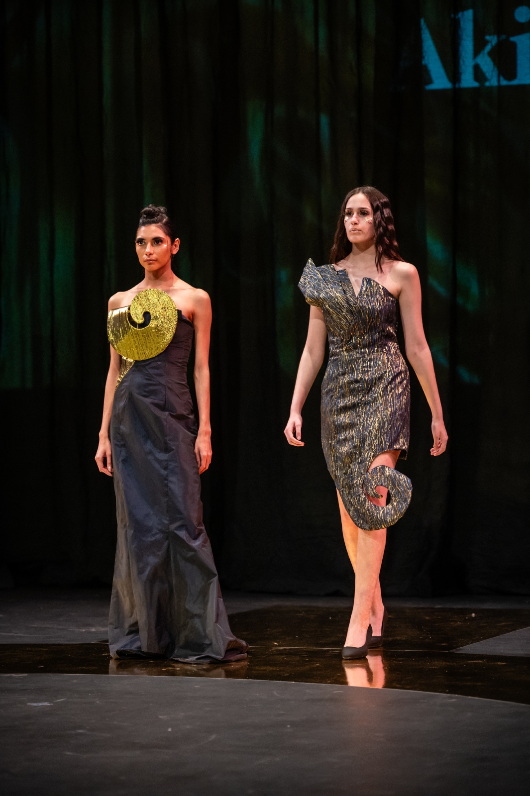 Two models join each other on the runway, wearing dark gray dresses with bright details