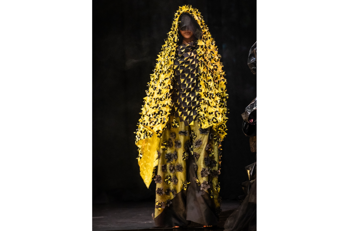 A model wearing a black and chartreuse garment with a large piece of textured fabric draped over the head, and a long skirt and pants