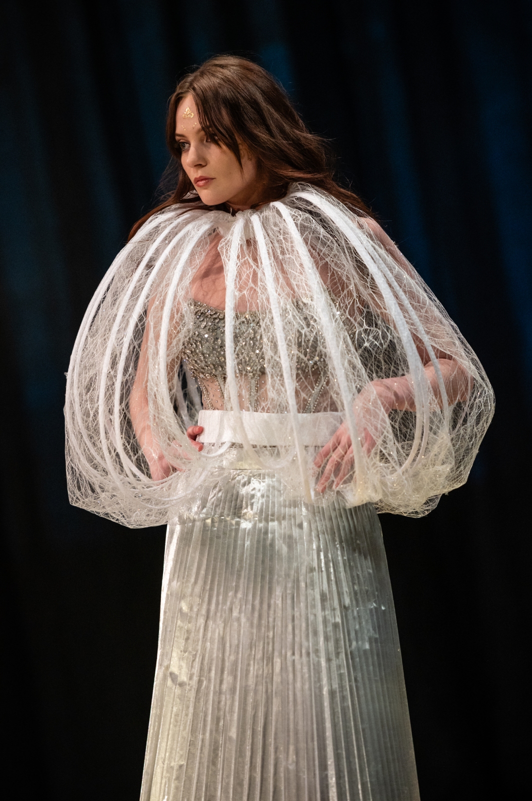 A model poses in a long white skirt with a curved, semi-sheer lace cover over her upper body