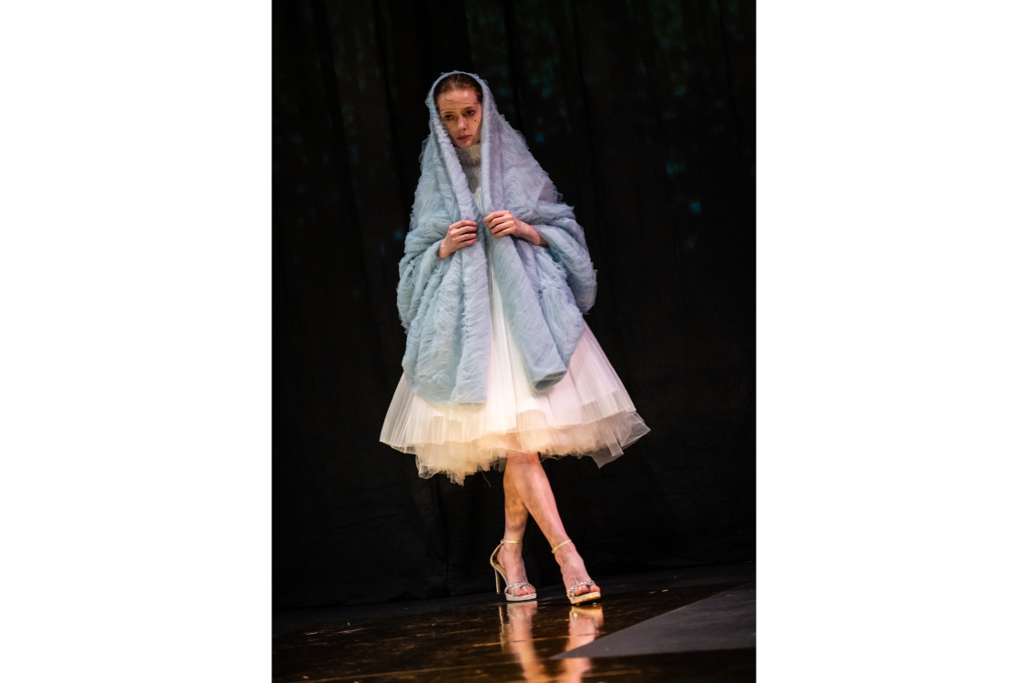 A model walks down the runway in a white, tulle ruffled skirt and a pastel blue shawl draped over her head and shoulders