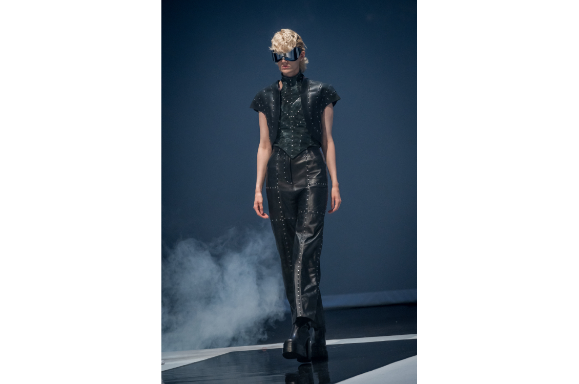 Runway model in black studded leather vest and pants with futuristic sunglasses.