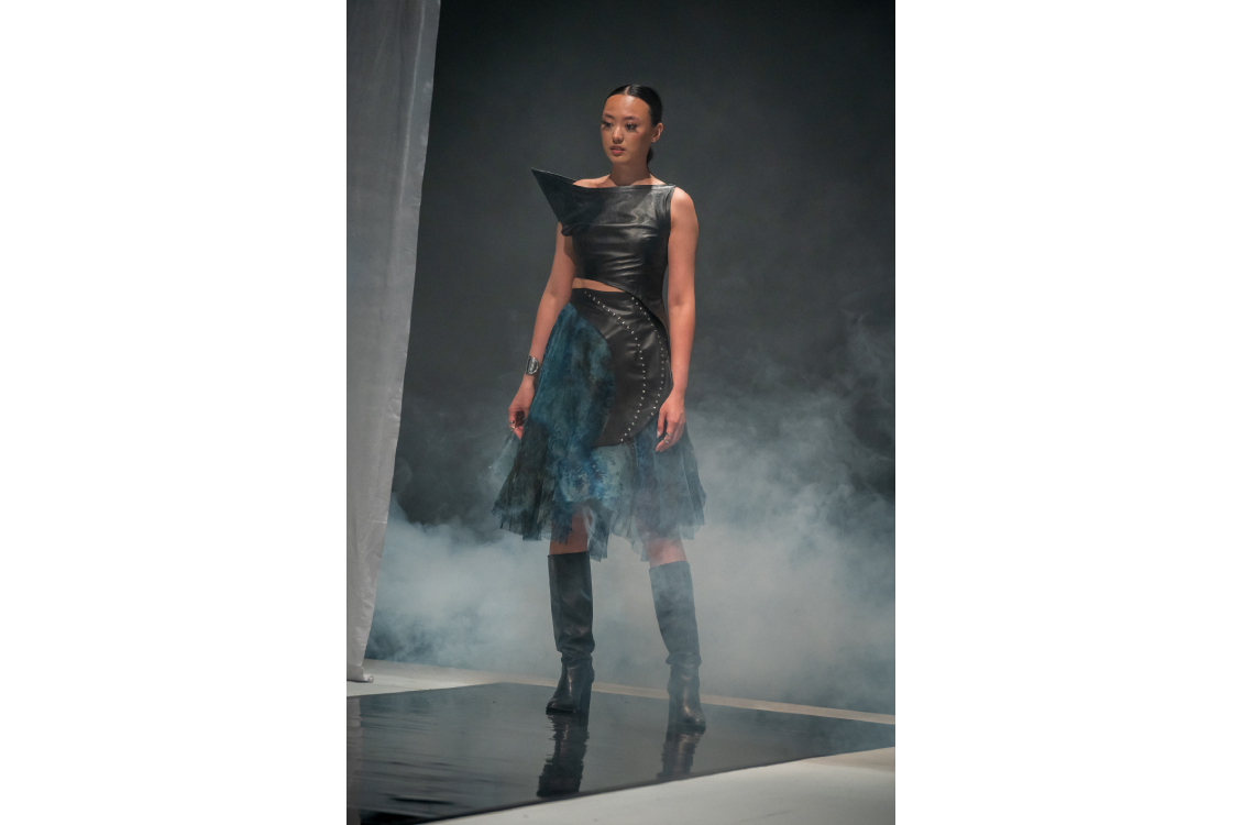 Runway model wearing an asymmetric black leather top and blue textured skirt.