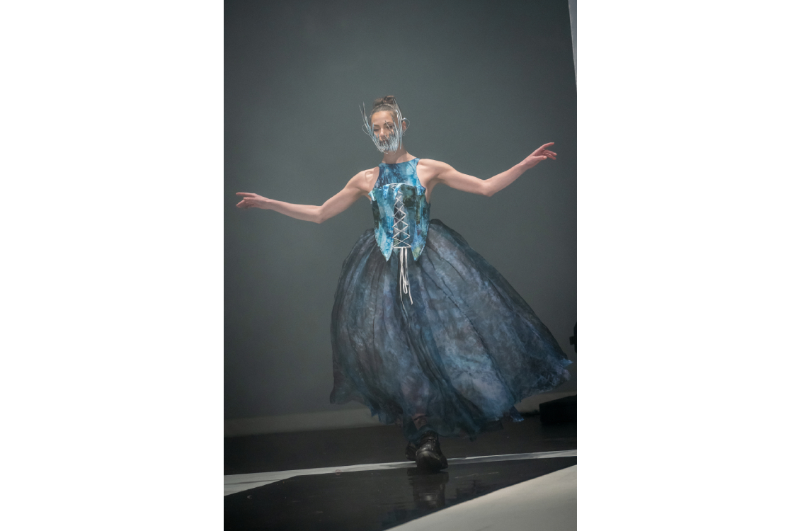 Runway model in a blue structured top and voluminous skirt with a sculptural face accessory.