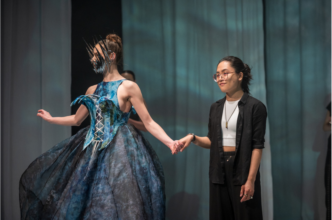 Designer Kaine Bautista stands on stage beside a model wearing her design