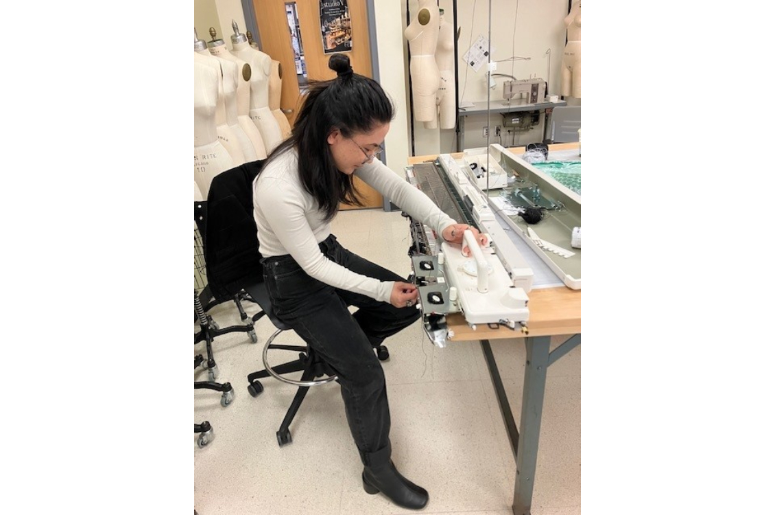 Kaine sits and works on industrial knitting machine