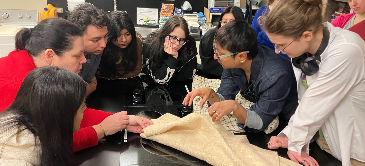 A professor, standing in the center, guides a group of students as they examine textile materials in a laboratory setting.