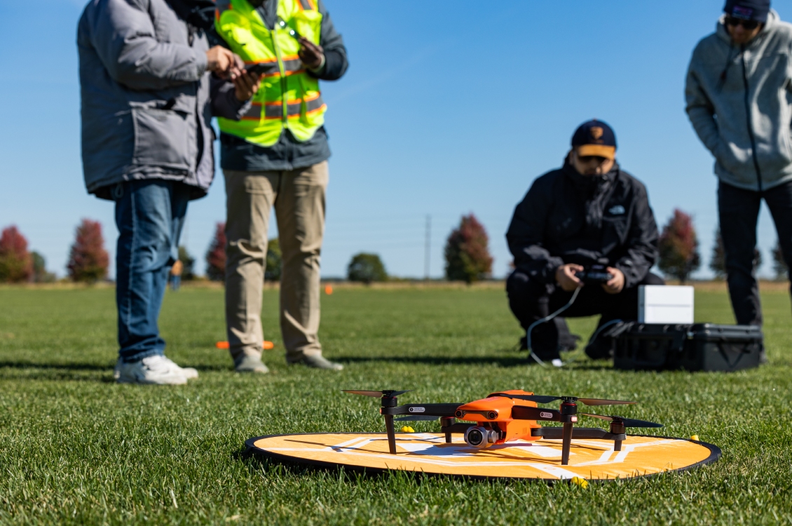 A student holding a remote control sits at a distance from the drone and landing pad on the ground.